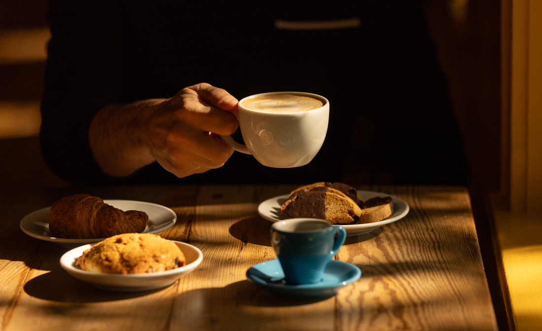 A cappuccino lifted over a café table set with chocolate-dipped biscotti, an espresso, and fresh pastries