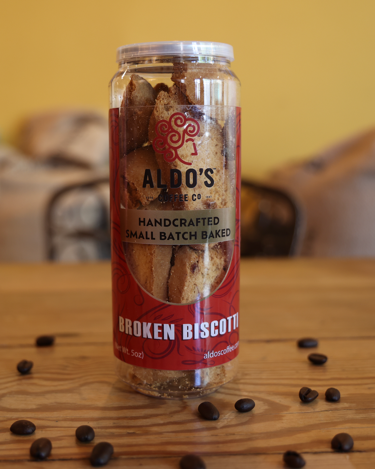 A clear plastic jar of Aldo's Coffee Co. Broken Biscotti cookies sits on a wooden table with scattered coffee beans.