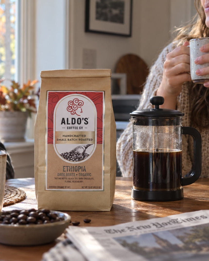 A bag of Aldo's Espresso coffee stands on a wooden table in a cozy kitchen setting, with a person on the left holding a mug and a person on the right holding a cup. A French press, mugs, coffee beans, and a newspaper are also visible.