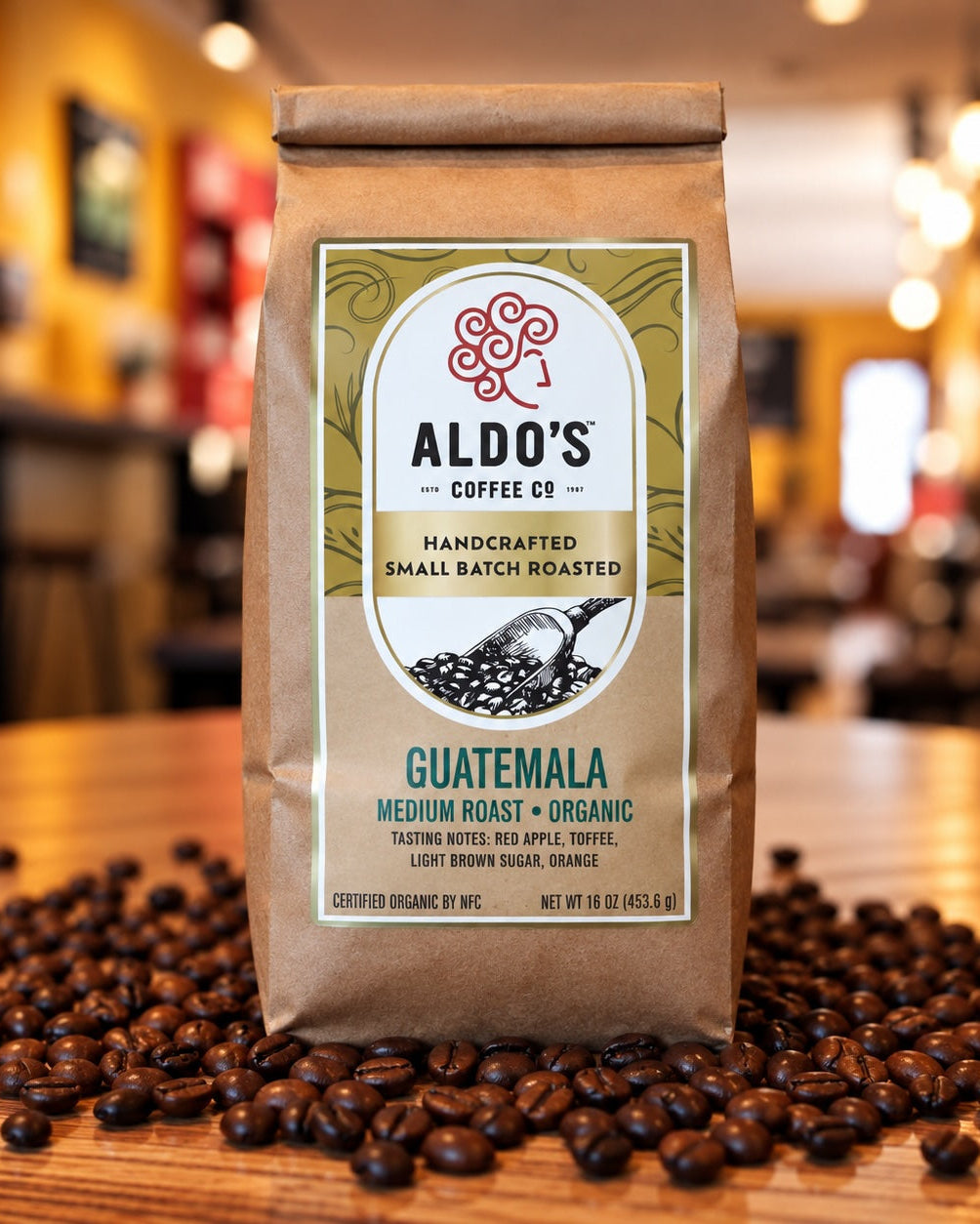 A bag of Aldo's Coffee Co. Guatemala medium roast organic coffee sits on a wooden table surrounded by scattered coffee beans, with a warm, blurred cafe interior in the background.
