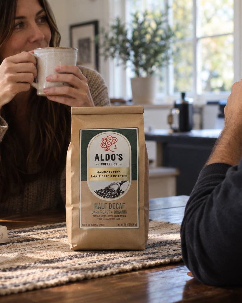 A bag of Aldo's Organic Half Decaf coffee sits on a wooden table between two people holding mugs in a cozy, sunlit kitchen setting.