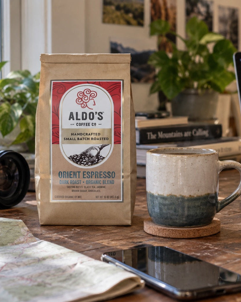 A bag of Aldo's Coffee Co. Orient Espresso sits on a wooden desk in a cozy home office setting, surrounded by a ceramic mug, laptop, camera, and a person writing in a notebook.