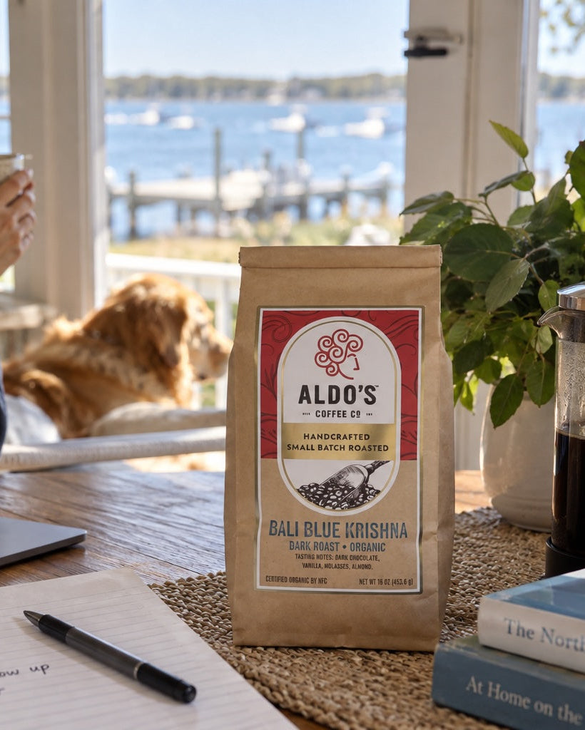 A bag of Aldo's Coffee Co. Ball Blue Grispa coffee sits on a wooden table in a cozy home office setting, with a woman working on a laptop and a dog lying nearby, with a French press and books in the foreground.