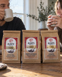 Three bags of Aldo's Coffee Roasted beans on a wooden table, with a man and woman holding mugs in the background in a cozy kitchen setting.