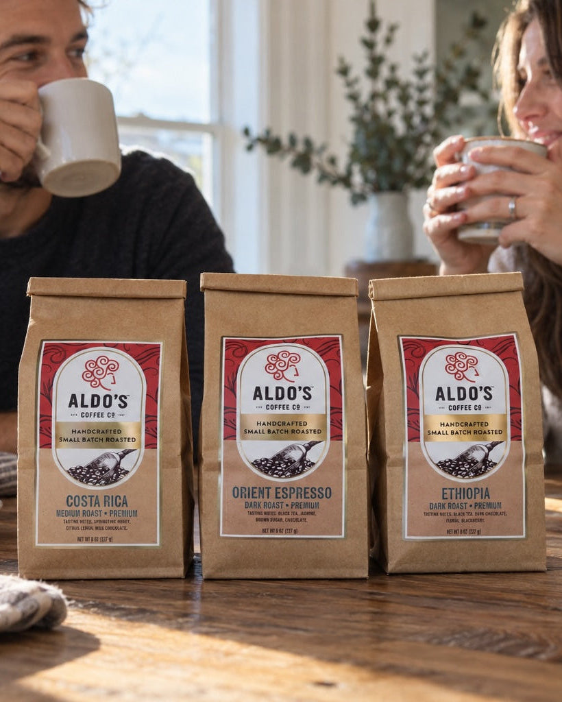 Three bags of Aldo's Coffee Roasted beans on a wooden table, with a man and woman holding mugs in the background in a cozy kitchen setting.